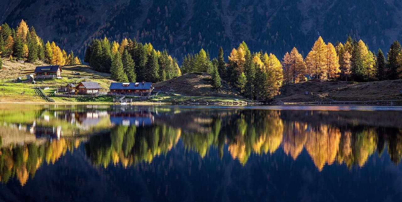 Landschaftsbild. 3 Häuser am See und im Hintergrund ein herbstlicher Wald.