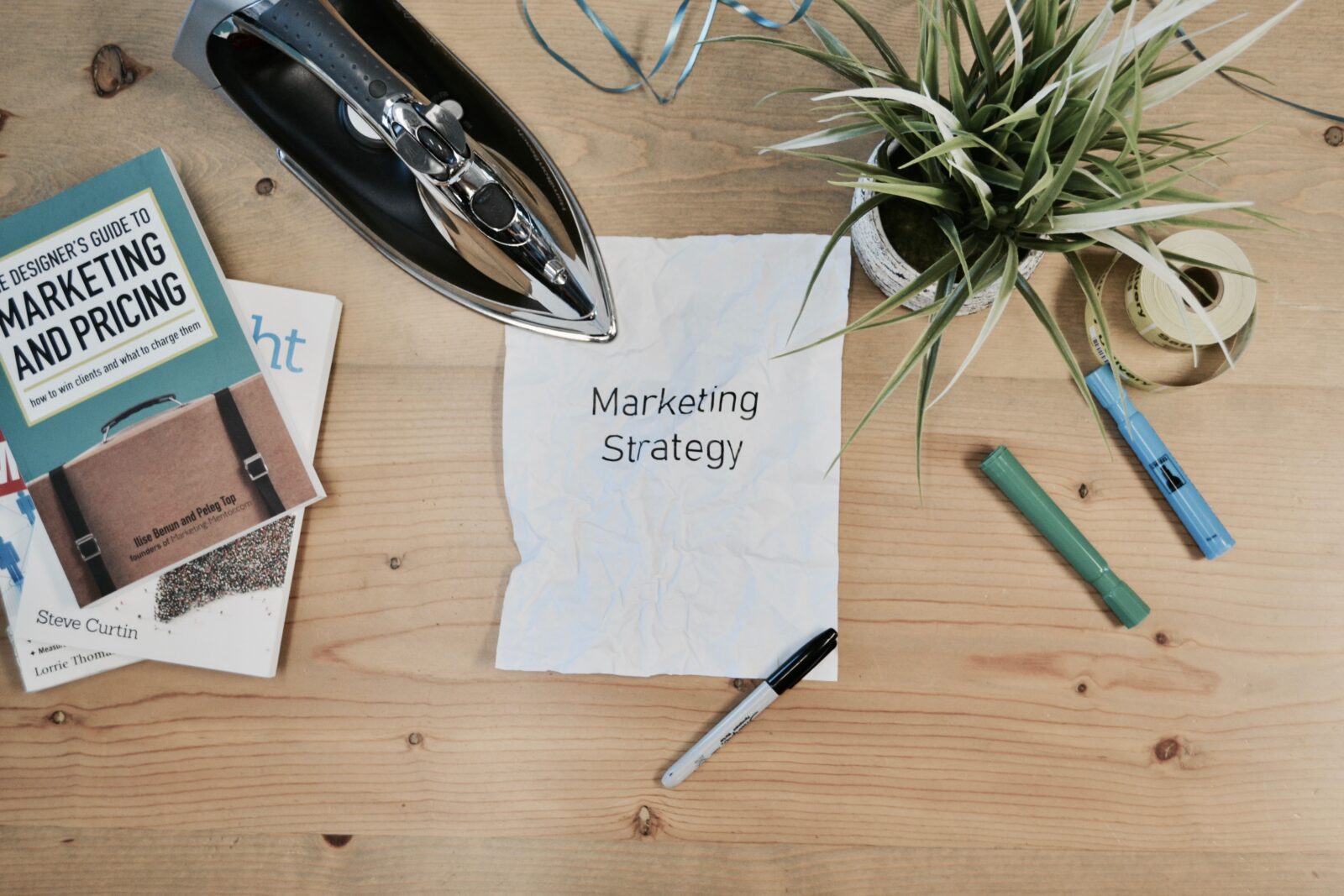 Tisch mit STiften, Büchern und einer Notiz. Auf der Notiz steht Marketing Strategy. Das ist die englische Bezeichnung ür Marketingplan oder Salesplan.